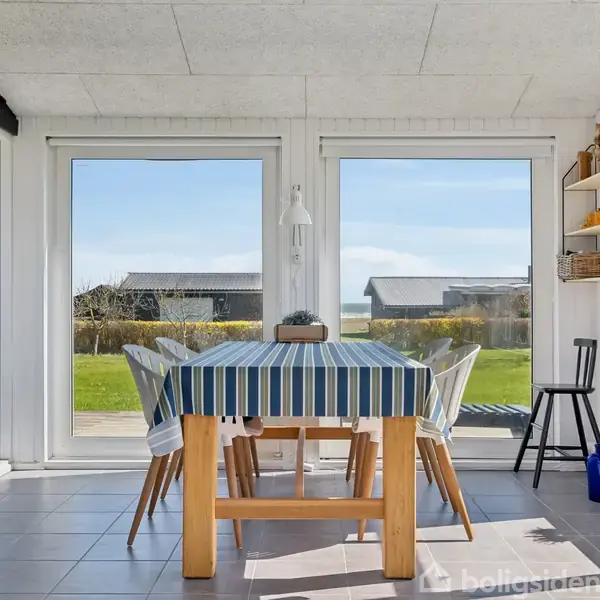Spisebord med stribet dug, omgivet af stole står i et lyst rum med store vinduer. Udsigt over grøn have og udendørs bygninger. Hylder med pynt ses til højre.