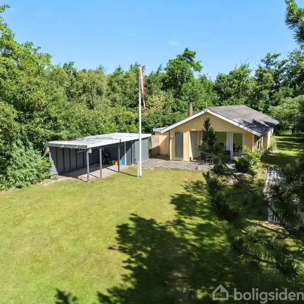 Huset står stille i en grøn have omgivet af træer med en carport ved siden af.