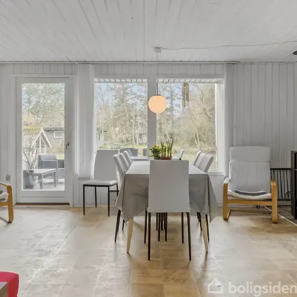 Et moderne spisebord med seks stole står i et lyst rum med store vinduer og en brændeovn i hjørnet. To stole med puder flankerer spiseområdet.