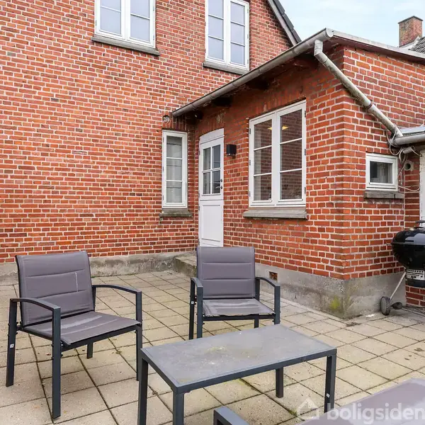 Rød murstensbygning med baggårdsmiljø. Tre sorte havestole og et bord står på en flisebelagt terrasse. Der er en sort grill i hjørnet ved siden af huset med hvide vinduer og dør.