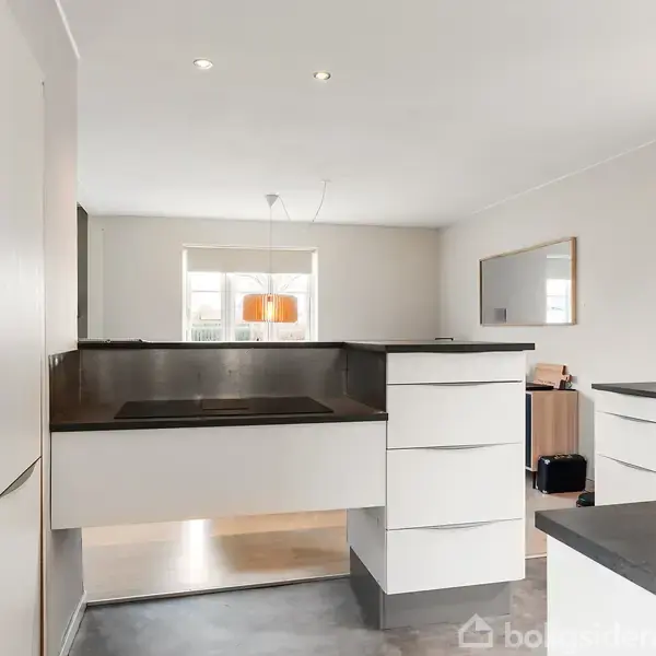 Elegante, moderne køkken med hvide skabe og integrerede apparater. En central kogeø dominerer rummet, mens naturligt lys strømmer ind fra et stort vindue. Enkel indretning i et lyst rum.