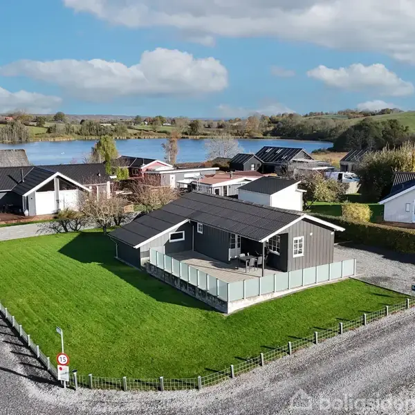 Gråt hus med terrasse står på en grøn græsplæne omgivet af grusveje i en stille forstad. Baggrunden viser en sø og flere huse. Der står en bil parkeret nær huset.