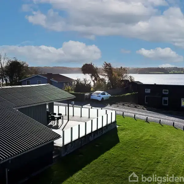 Et hus med sort tag står stille ved en stor terrasse. I baggrunden ses en sø omgivet af træer og en bakket landskab under en delvist skyet himmel.