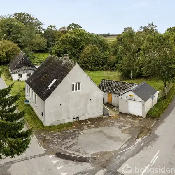 Hus med skråt tag og hvide vægge står ved en vej. Det er omgivet af træer og landskab, og der er et skur ved siden af.