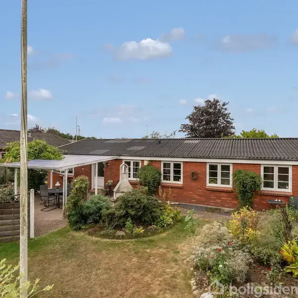 En etplans rødstenshus står foran en velplejet have med buske og blomster. Ved siden af huset er en terrasse med havemøbler. I baggrunden ses flere huse og træer.