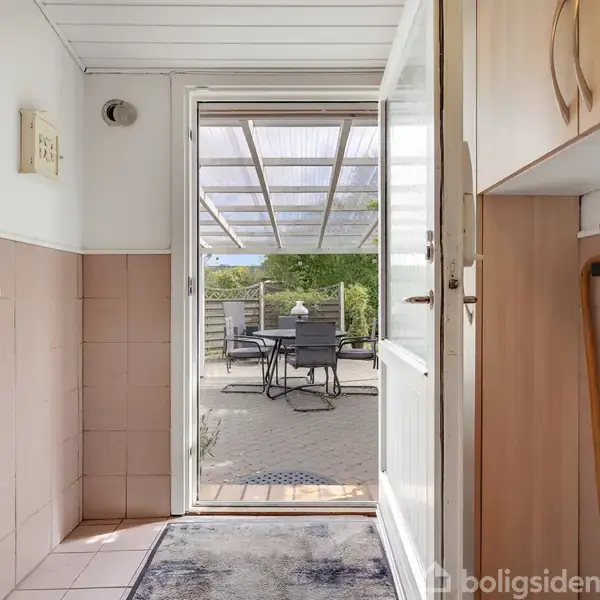 Dør åbner ud mod overdækket terrasse med spisebord og stole i baghaven. Indgangen har hvide vægge og lyserøde fliser, med en højre side opbevaringsplads.