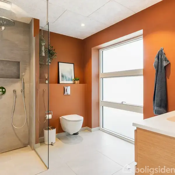 Brusekabine med glasvæg, håndbruser og regnbruser; ved siden af hænger et håndklæde. Kontekst: moderne badeværelse med terracotta-væg, toilet, vindue og vask med skabsplads.