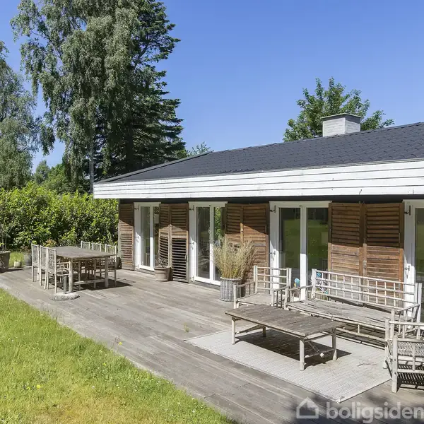 Stue i lys, minimalistisk stil med hvid sofa, sofabord og fladskærms-TV. Rummet har store vinduer med grøn udsigt udenfor, samt lys trægulv og en stor terrakottakrukke.