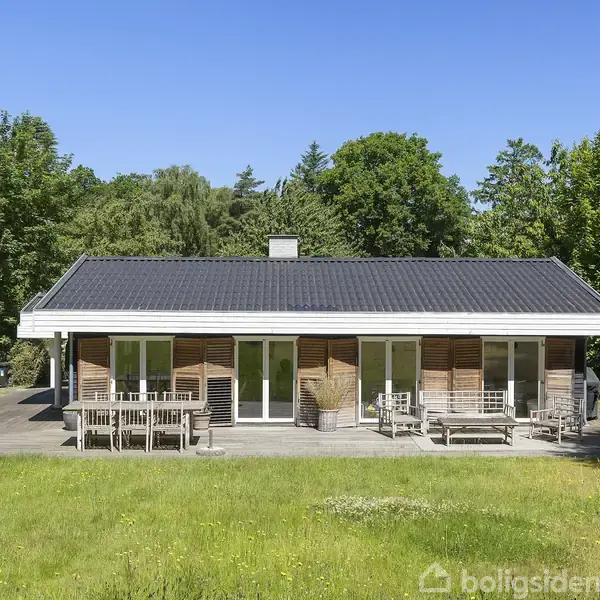 Stue med hvidmalede vægge og loft. En stor sofa står til højre, og et spisebord er i baggrunden. Store vinduer giver udsigt til en have. En bog ligger på et bord.