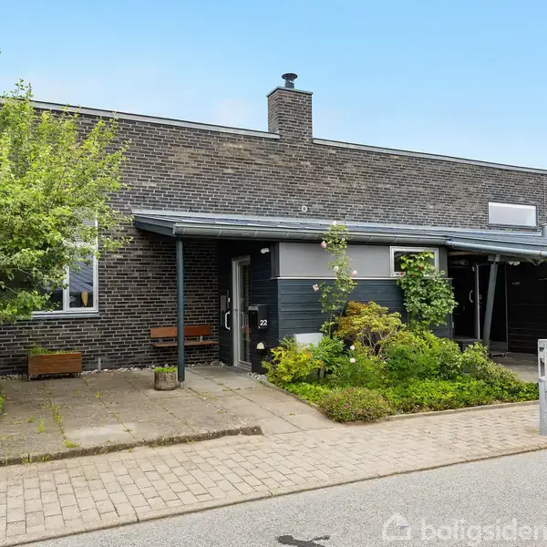 Et moderne rækkehus med fladt tag og mørke mursten som hovedmateriale. Foran huset er der et grusbelagt fortov og nogle grønne buskads. En velplejet forhave pryder indgangen.