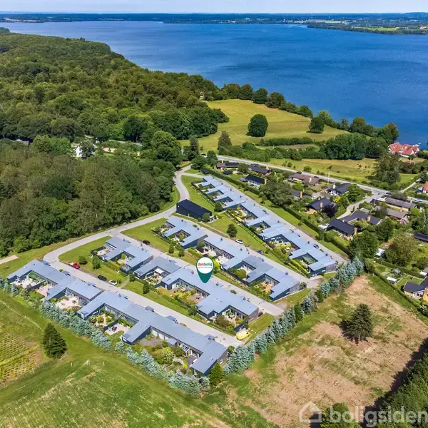 Boligkompleks fra luften med rækkehuse arrangeret i zigzag-mønster. Beliggende nær kystlinje og omgivet af træer og landbrugsjord, med udsigt over en stor sø og skovområde.