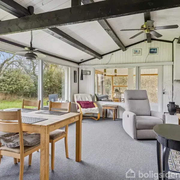 Spisebord med stole står i en lys stue med store vinduer. I baggrunden ses en sofa og lænestol. Udenfor er en frodig have. Loftet har synlige bjælker og en loftsventilator.