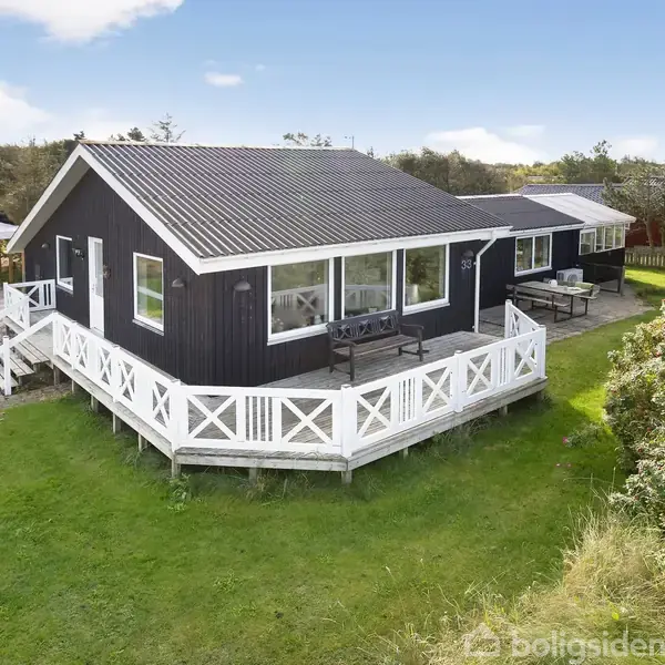 Et sort træhus med hvide vinduer og stor terrasse står på en grøn græsplæne. Terrassen har hvidt rækværk, og der er havemøbler. Omkring huset er der buske og træer.