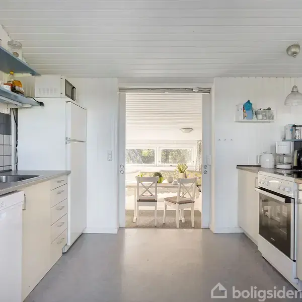 Et lyst køkken med hvidt interiør. Til venstre er der en vask og en opvaskemaskine. I midten er en åbning til en spisestue med hvide stole. Tekst på billedet: "boligsiden".