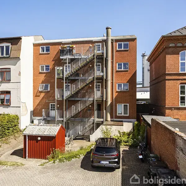 Bygning med udendørs metaltrappe, står i solen mellem to andre murstensbygninger. En parkeret bil og lille rød skur ses i gårdspladsen omgivet af brostensbelægning.