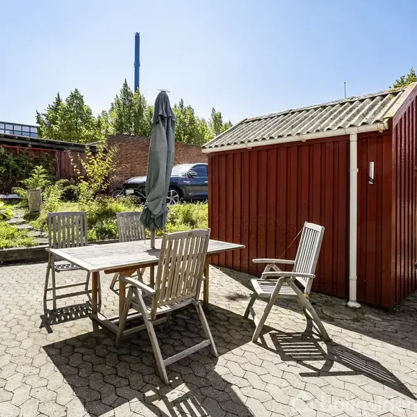 Rød skur står på en flisebelagt terrasse. Ved siden af er der et bord med fire stole og en grøn parasol. Baggrunden har en mur og træer under en klar blå himmel.
