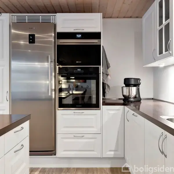 Et køkken med hvidt skabe og en stål køleskab til venstre. Ovne er indbygget i midten, med en køkkenmaskine ved siden af. Træloftet giver rummet en varm atmosfære.