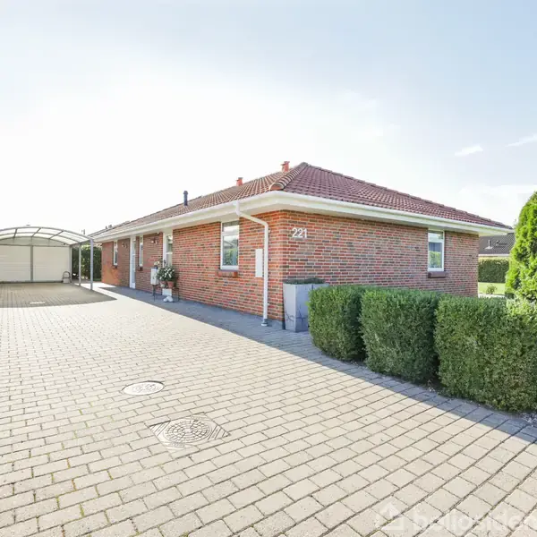 En rød murstensvilla med rødt tegltag står på en flisebelagt indkørsel. Foran huset er der grønne buske. Nummer "22" er synligt på ydervæggen ved døren.