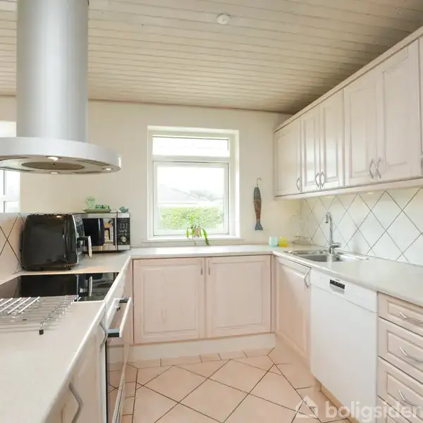 Køkken med hvidt interiør, indeholdende grå emhætte over komfur og ovn. Hvidlakerede skabe fylder væggene. Et vindue med lysindfald og planter. Kaffemaskine og brødrister på bordet.