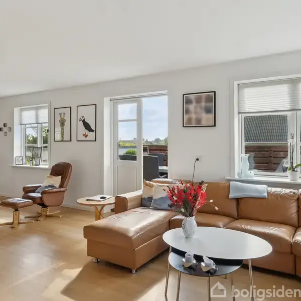 Brun hjørnesofa med farverig buket på rundt sofabord, står i en lys stue med store vinduer og lysindfald. Billeder hænger på væggen; spisebord og lænestol ses i baggrunden.