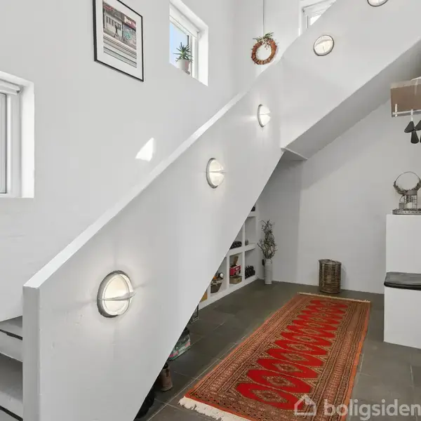 Trappeopgang med indbyggede lys går opad. Rød orientalsk tæppe ligger på gulvet. Vægge er hvide med små vinduer. Et lille bord med dekorative genstande står i hjørnet.