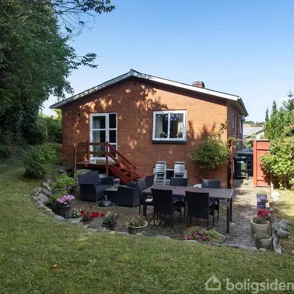 Murstenshus med terrassebord og stole i haven, omgivet af græsplæne og træer. En række blomsterpotter dekorerer området. Stolen er tæt på huset, der har to vinduer.