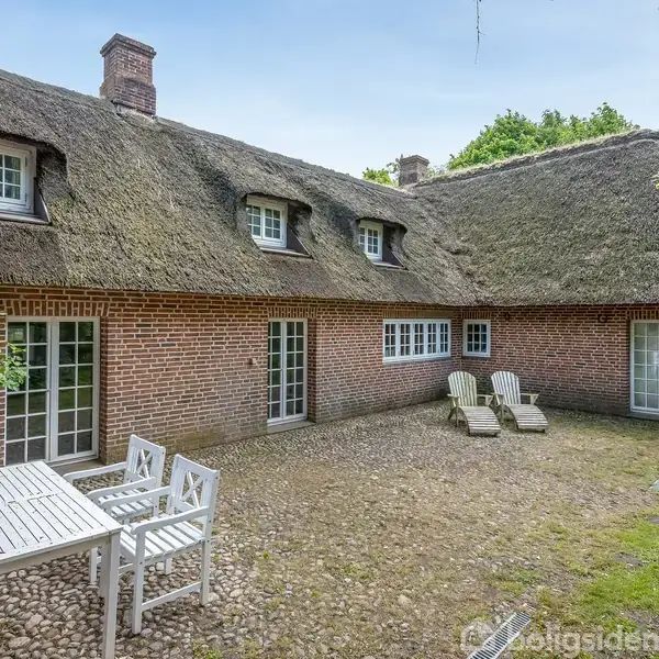 Et rundt bord med blomster og bakværk står i en entré med sort/hvid-ternet gulv. En lysekrone hænger over bordet. En antik ur står ved siden af en trappe.