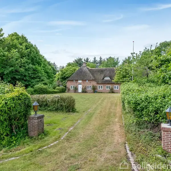 Et lyst køkken med sort-hvidt ternet gulv. Et stort køkkenbord med træbordplade står centralt. Skabe er fyldt med tallerkener og glas. En rød standerur står i hjørnet.