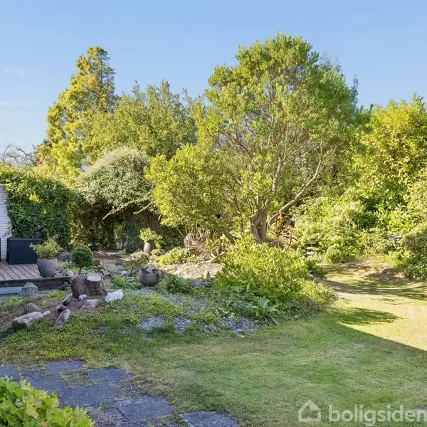 Terrasse med udendørs siddepladser ved siden af et hus; omgivet af en frodig have med græsplæne og buske. Flisebelagt område fører til en gangsti.