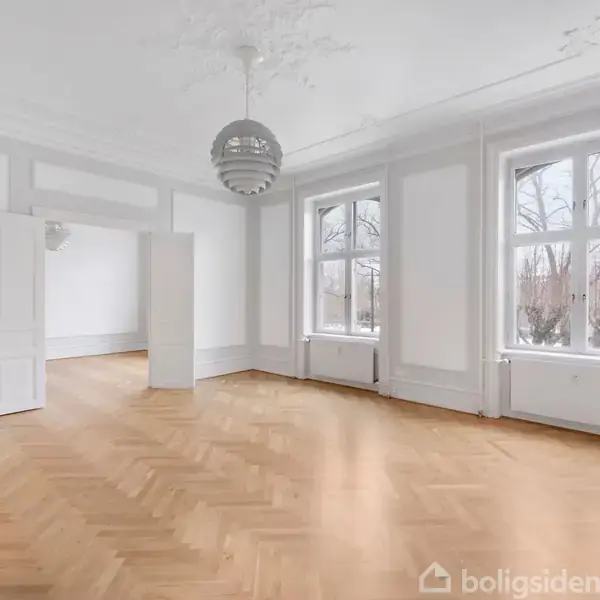 Et stort, tomt rum med trægulv og højt til loftet. En dekorativ loftslampe hænger centralt. To store vinduer giver udsigt til træer udenfor. Væggene er lyse, og der er to åbne døre.