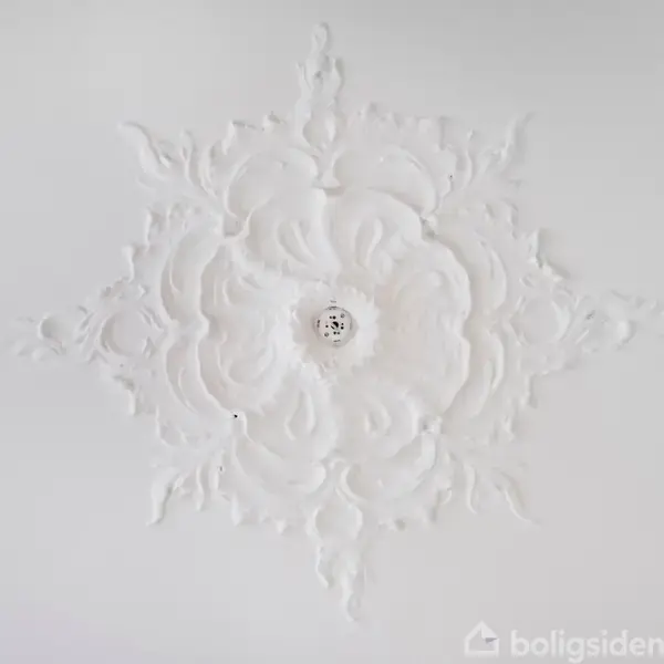 Loftsroset udført i detaljeret hvid stuk i et geometrisk mønster med en lille rund lampe i midten. Rosetten er centreret på en glat hvid loftsoverflade.