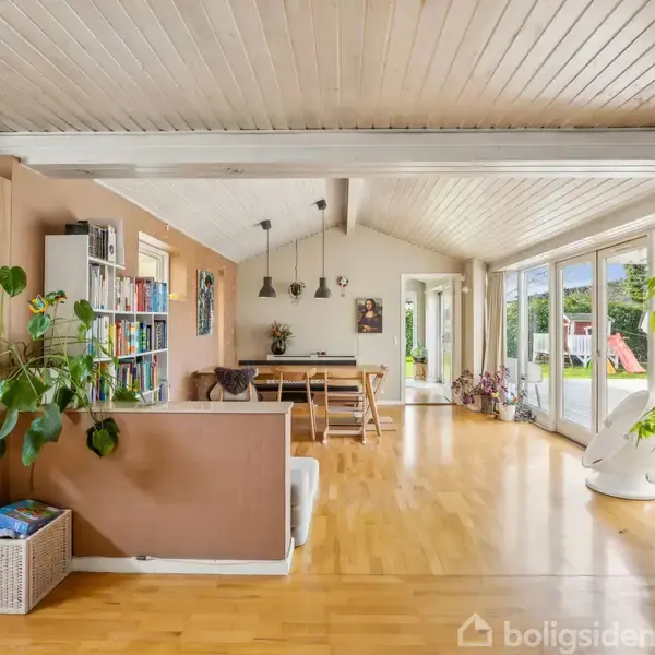 Stue med trægulv dominerer rummet; bøger og planter fylder reolerne. Et spisebord med stole står midt i rummet. Store vinduer giver udsigt til en have.