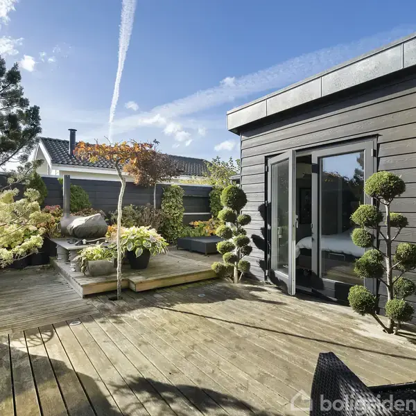 Terrasse med trædæk og moderne sorte møbler. Planter og buske pryder omgivelserne. Bygninger i skandinavisk stil omkranser området under en klar blå himmel.