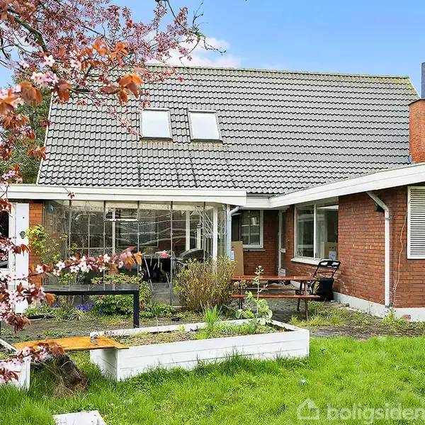 Rødt murstenshus med skråt gråt tegltag står i en have med grønne græsplæner. Blomstrende træ og planter omgiver en terrasse med borde og stole i sollys.