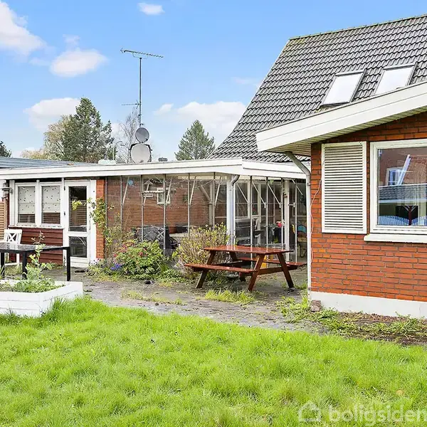 Rødt teglhus med hvide vinduer står stille i en grøn have med græs og en træterrasse. Der er et havebord med stole og et drivhus ved siden af huset.