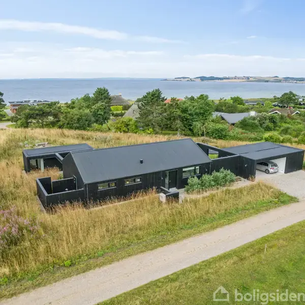 Et sort, moderne hus ligger omgivet af højt græs nær en grusvej, med udsigt til en kystlinje i baggrunden. Omkringliggende huse og træer skaber en rolig atmosfære.