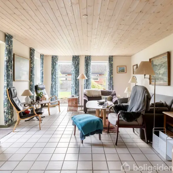 Stue med sofaer og lænestole placeret omkring et sofabord. Store vinduer med blå gardiner giver udsigt til en have. Flisegulv og træloft kompletterer den lyse indretning.
