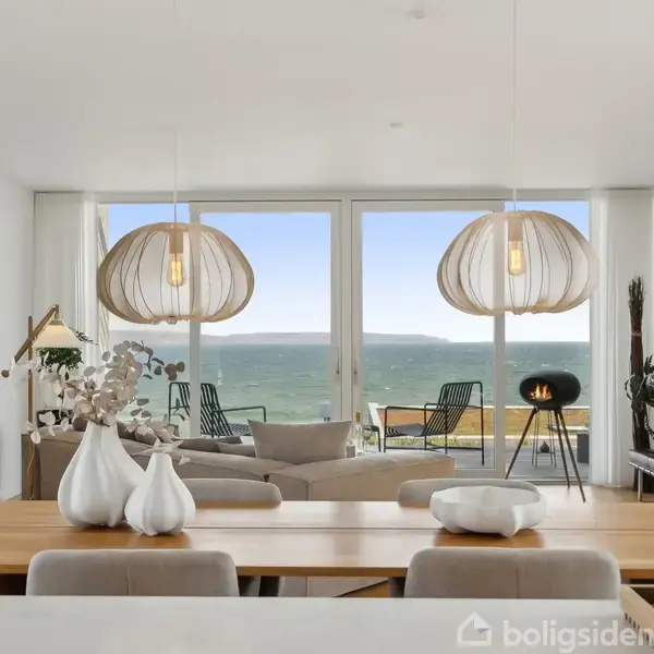 Spisebord er prydet med dekorative vaser. Bagved ses en stue med moderne belysningsarmaturer og en sofa. Store vinduer afslører en balkon med havudsigt og udendørs møbler.