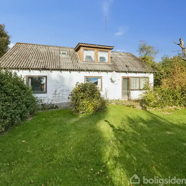 Hus med hvid facade står i en have med grøn græsplæne. Taget er gråt med ovenvinduer, omgivet af træer og buske under en klar blå himmel.