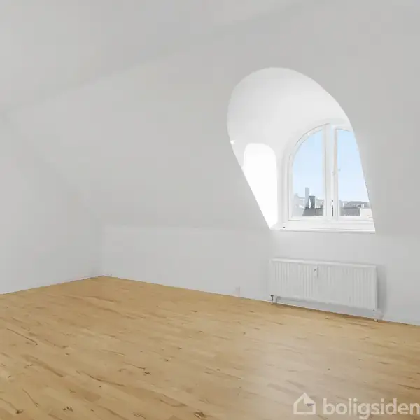 Et lyst, tomt værelse med trægulv har et buet vindue, der tillader dagslys at strømme ind. En radiator er placeret under vinduet, og rummet har hvide vægge.