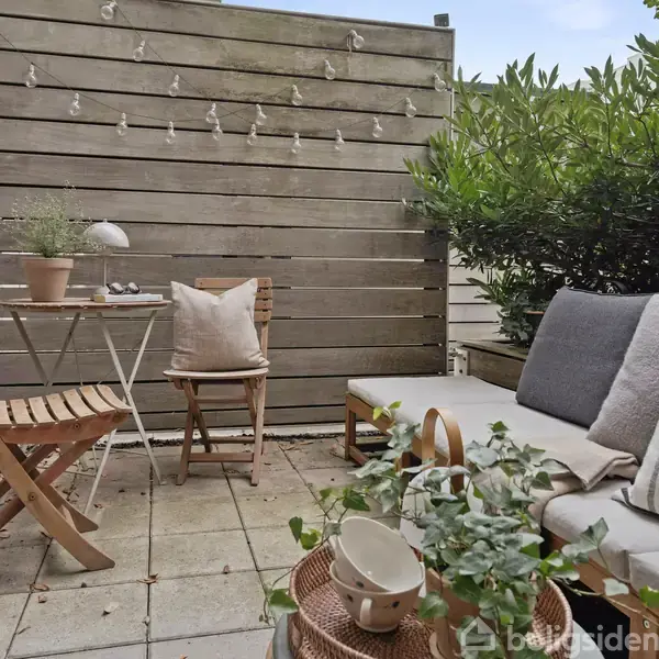 Terrasseindretning med træsidebord og stol med pude, placeret ved siden af en sofa med tæpper. Omkranset af grøn beplantning og afgrænset med trævæg dekoreret med lyskæde.