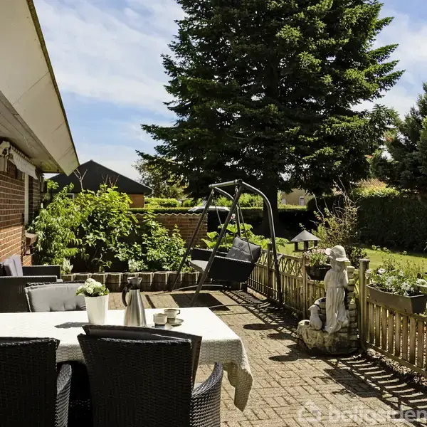 En terrasse med borde og kurvestole står ved et murstenshus. En gynge ses i baggrunden. Omgivet af planter og træer i en have.