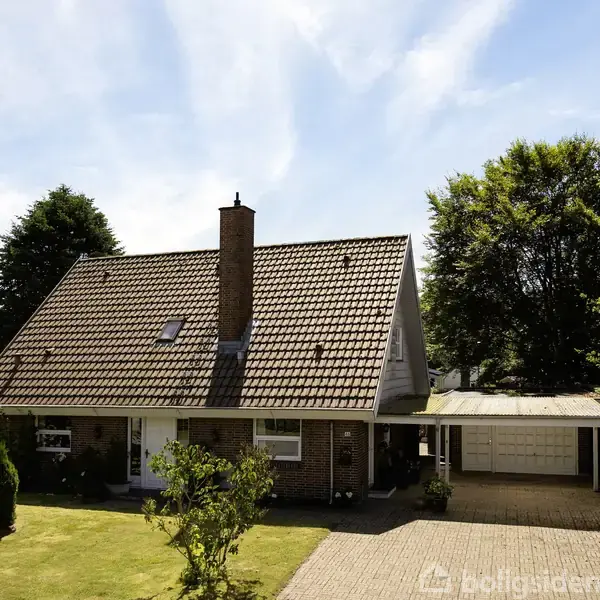 Et rødstenshus med spidst tag står stille i en velplejet have med grønne træer omkring. En garage er synlig til højre for huset.