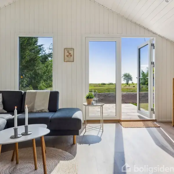 En stue med sofa og sofabord, der står ved siden af et åbent glasdørsparti. Udsigt til en terrasse og grønne områder udenfor i dagslys. Ingen tekst fundet.