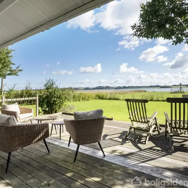 Terrassemøbler står på en overdækket træterrasse. Møblerne, to stole og to loungestole, vender ud mod en stor have med udsigt over en sø under en lys, skyet himmel.