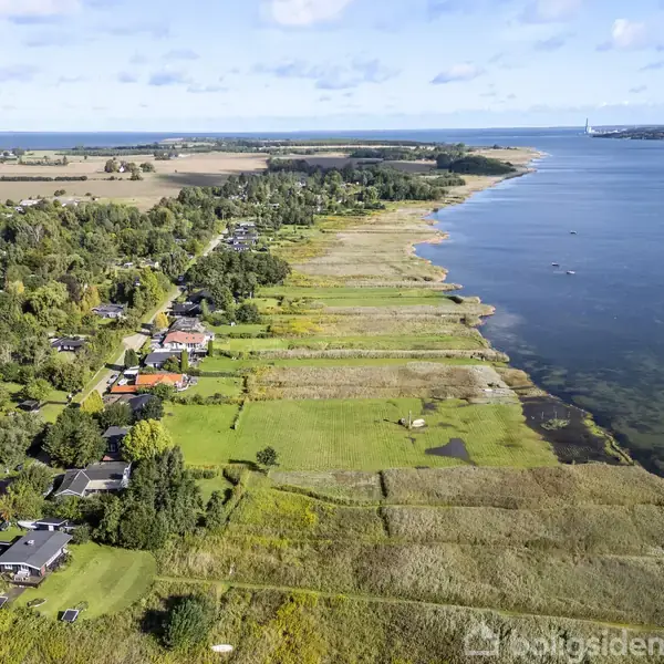 Kystlandskab set fra oven viser et grønt område med spredte huse langs en strandlinje ved en rolig sø. Området omgivet af åbne marker og skove i baggrunden.