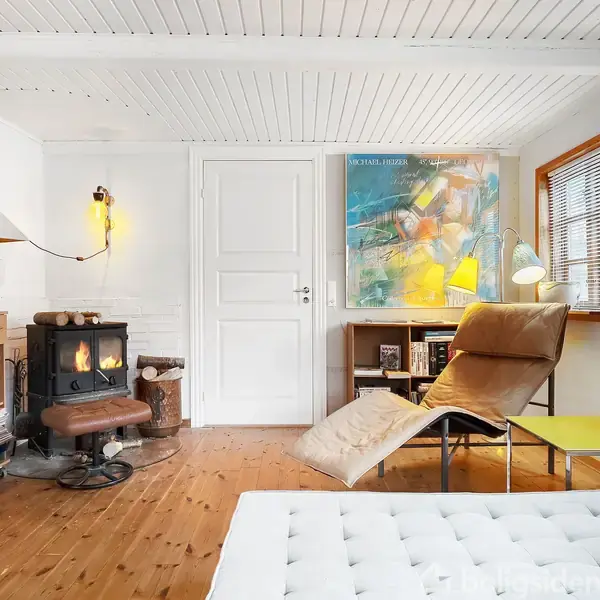 Pejs varmer stue med trægulve og hvide vægge. Loungestol ved gulvlampe og gult bord. Kunst pryder væggene, mens en reol og pejs fuldender den hyggelige atmosfære.