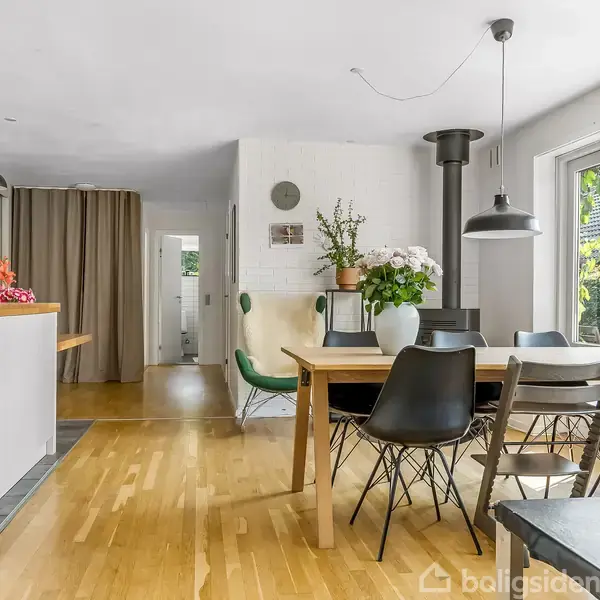 Spisebord med sorte stole står i lyse spiseområde. Plante og blomster på bordet, hængelamper over. Store vinduer til haven giver naturligt lys. Trægulv og moderne indretning.