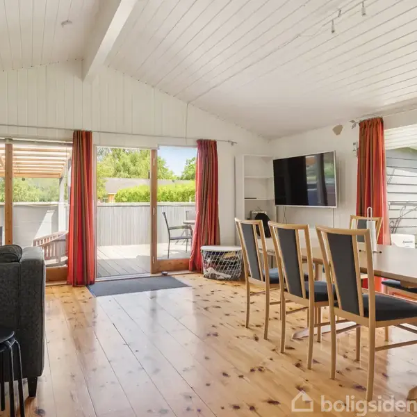 Stue med træmøbler, sofagruppe og balkonadgang gennem store glasdøre. Trægulv og hvidt loft, omgivet af vinduer med røde gardiner. Vægmonteret TV ved siden af spisebordet.