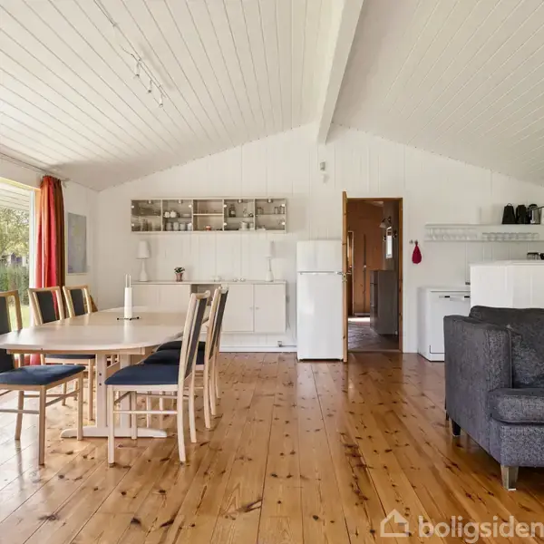 Lyse træmøbler står i et åbent køkken-alrum med spisebord og stole. Til venstre er store vinduer med udsigt til en grøn have. En grå sofa er placeret ved køkkenet.